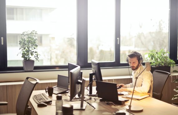 A man in a white shirt sitting at a desk with two computers.
